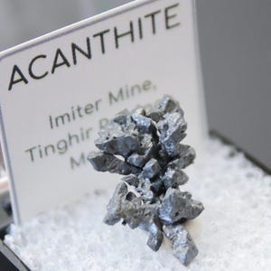 May include: Acanthite mineral specimen displayed in a black box with a white, textured interior. The mineral is a cluster of dark gray, metallic crystals. A white card with the word "ACANTHITE" and other text is in the background.