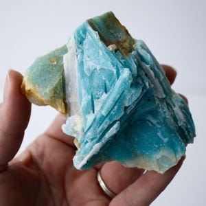 May include: A hand holds a vibrant, raw mineral specimen. The stone features a striking combination of colors, including a bright turquoise, pale green, and hints of brown. The crystal structure is visible, with sharp, angular formations.
