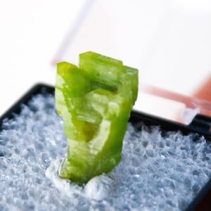 May include: A vibrant, lime-green crystal specimen displayed in a small, black, hinged display case. The crystal is set against a bed of light gray foam. The case is held open, revealing the crystal's unique shape and color.
