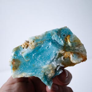 May include: A vibrant, raw gemstone with a striking blue hue, accented by areas of white and golden-brown. The mineral's rough texture and irregular shape are visible. The stone is held in a hand against a white background.