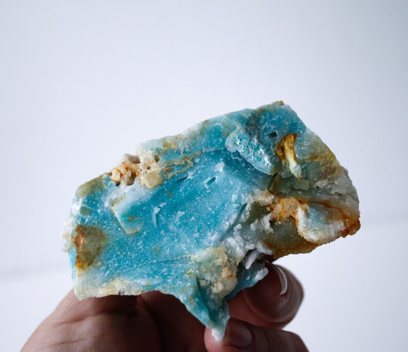 May include: A vibrant, raw gemstone with a striking blue hue, accented by areas of white and golden-brown. The mineral's rough texture and irregular shape are visible. The stone is held in a hand against a white background.
