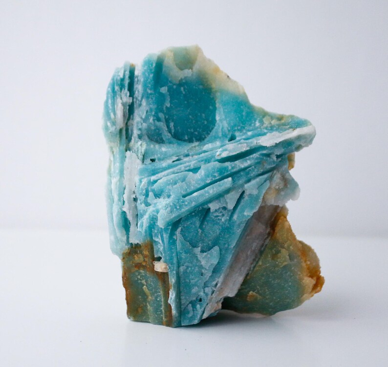 May include: A striking, raw amazonite gemstone with a vibrant turquoise hue. The stone features a layered, textured surface with areas of white, brown, and green. This natural crystal is a unique piece for collectors or those interested in minerals.
