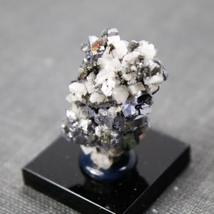 May include: A cluster of gray, white, and black crystals on a black display stand. The crystals are arranged in a random pattern and have a variety of shapes and sizes.
