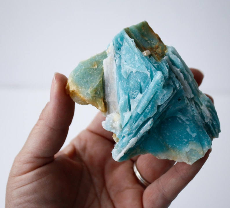 May include: A hand holds a vibrant, raw mineral specimen. The stone features a striking combination of colors, including a bright turquoise, pale green, and hints of brown. The crystal structure is visible, with sharp, angular formations.