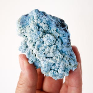 May include: A hand holding a light blue mineral specimen. The mineral has a bubbly, textured surface with areas of darker blue and black. The overall shape is irregular and organic, with a crystalline appearance.