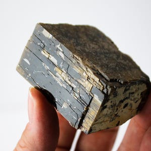 May include: A dark, rectangular mineral specimen with a layered appearance. The stone features a mix of dark gray, black, and golden-brown hues. The rough edges and natural texture suggest a raw, unpolished state, highlighting its geological origins.