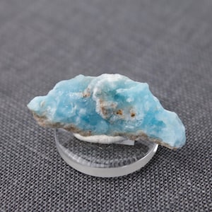 May include: A light blue celestite crystal with a rough, textured surface. The crystal is on a clear glass display stand.