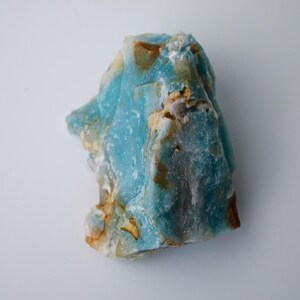 May include: A rough-cut, natural stone with a striking blue hue, accented by patches of tan and brown. The mineral's surface has a textured appearance, showcasing its raw, unpolished state. The stone is set against a plain white background.