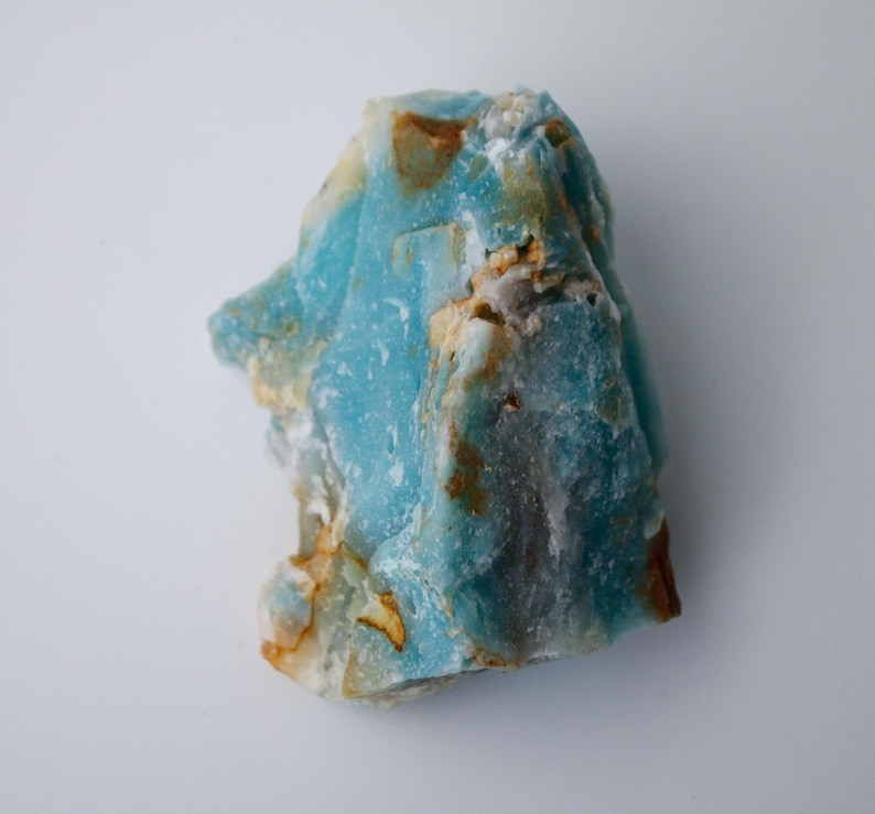 May include: A rough-cut, natural stone with a striking blue hue, accented by patches of tan and brown. The mineral's surface has a textured appearance, showcasing its raw, unpolished state. The stone is set against a plain white background.