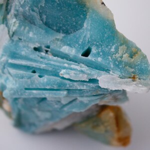 May include: A close-up of a rough, natural mineral specimen. The stone is primarily a vibrant turquoise blue with areas of white and tan. The texture is layered and crystalline, with visible crevices and holes. The overall shape is irregular and organic.