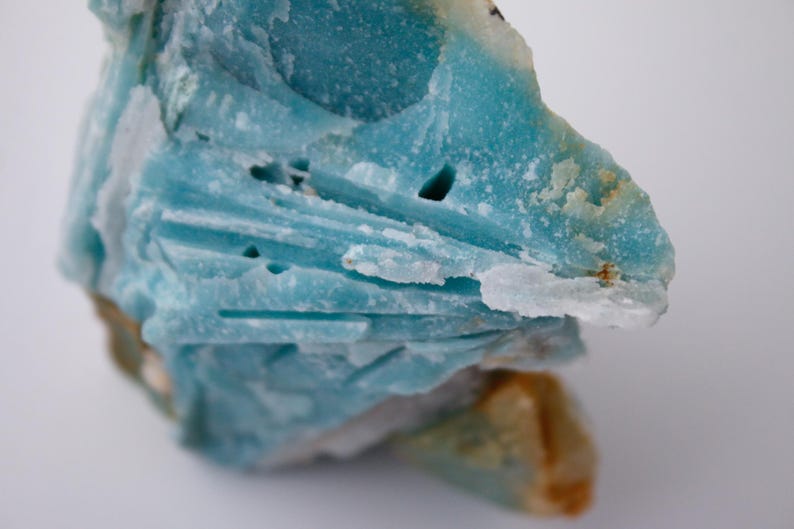 May include: A close-up of a rough, natural mineral specimen. The stone is primarily a vibrant turquoise blue with areas of white and tan. The texture is layered and crystalline, with visible crevices and holes. The overall shape is irregular and organic.