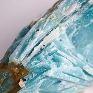 May include: Close-up of a rough, natural mineral specimen. The stone features layers of light blue and white crystalline structures with areas of brown and yellow hues. The surface has a textured, uneven appearance.