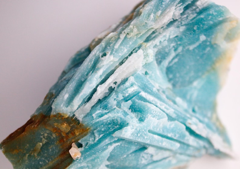 May include: Close-up of a rough, natural mineral specimen. The stone features layers of light blue and white crystalline structures with areas of brown and yellow hues. The surface has a textured, uneven appearance.