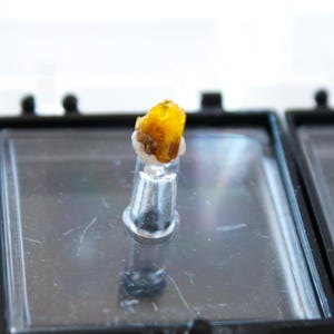 May include: A small, yellow, translucent crystal specimen mounted on a clear plastic base. The crystal is in a small, black display case.