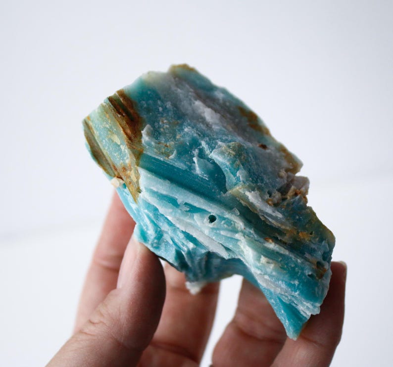 May include: A piece of raw, natural Amazonite gemstone. The stone displays a vibrant turquoise color with streaks of brown and white. The rough texture and unique patterns make it a striking specimen for collectors or jewelry makers.
