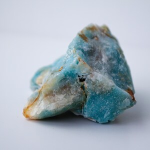 May include: A close-up of a rough-cut gemstone with a mix of blue, white, and tan colors. The stone has a textured surface and irregular shape, with areas of deep turquoise and lighter, opaque sections. The overall appearance is natural and earthy.
