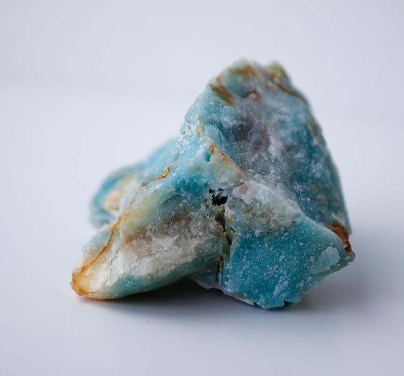 May include: A close-up of a rough-cut gemstone with a mix of blue, white, and tan colors. The stone has a textured surface and irregular shape, with areas of deep turquoise and lighter, opaque sections. The overall appearance is natural and earthy.