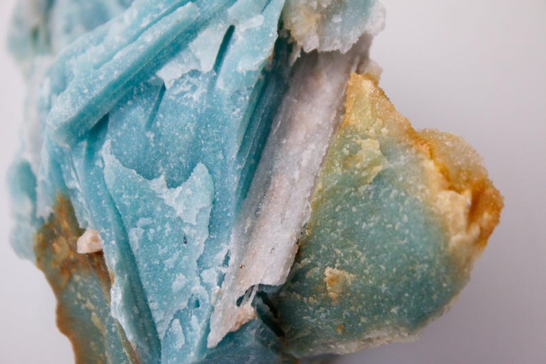 May include: Close-up of a mineral specimen featuring layered, crystalline structures. The dominant colors are shades of blue, with areas of white and touches of orange and brown. The texture appears rough and natural.