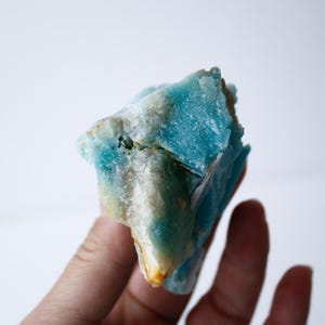May include: A close-up of a rough, natural stone with a striking blue hue, accented by areas of white and hints of yellow. The stone's surface is textured and irregular, showcasing its raw, unpolished state. The stone is held in a hand.