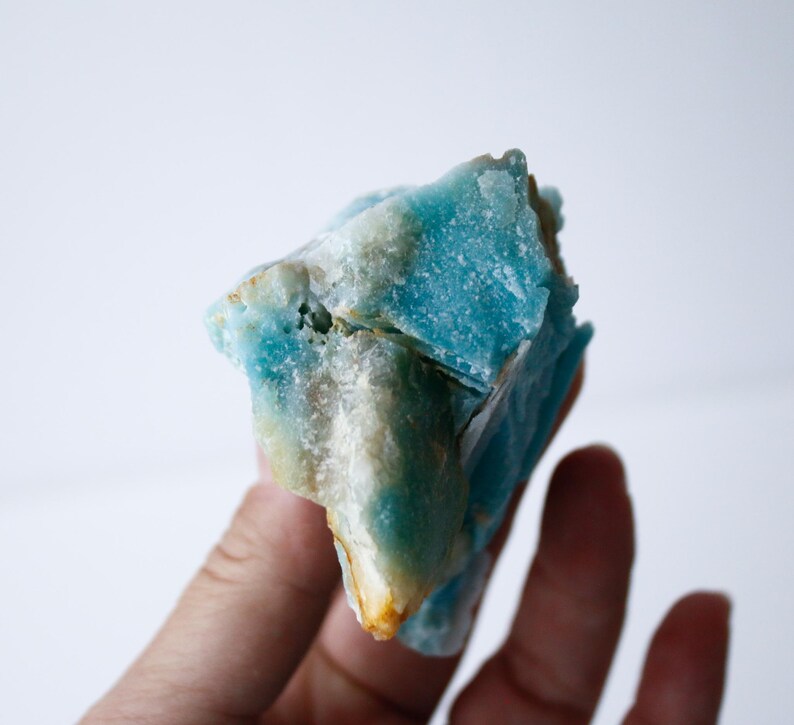 May include: A close-up of a rough, natural stone with a striking blue hue, accented by areas of white and hints of yellow. The stone's surface is textured and irregular, showcasing its raw, unpolished state. The stone is held in a hand.