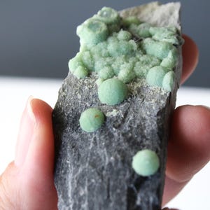 May include: A raw mineral specimen featuring a dark gray, jagged rock matrix embedded with clusters of small, spherical, light green crystals. The surface texture of the stone is rough and uneven, contrasting with the smooth, rounded appearance of the green mineral formations scattered across the top and sides.