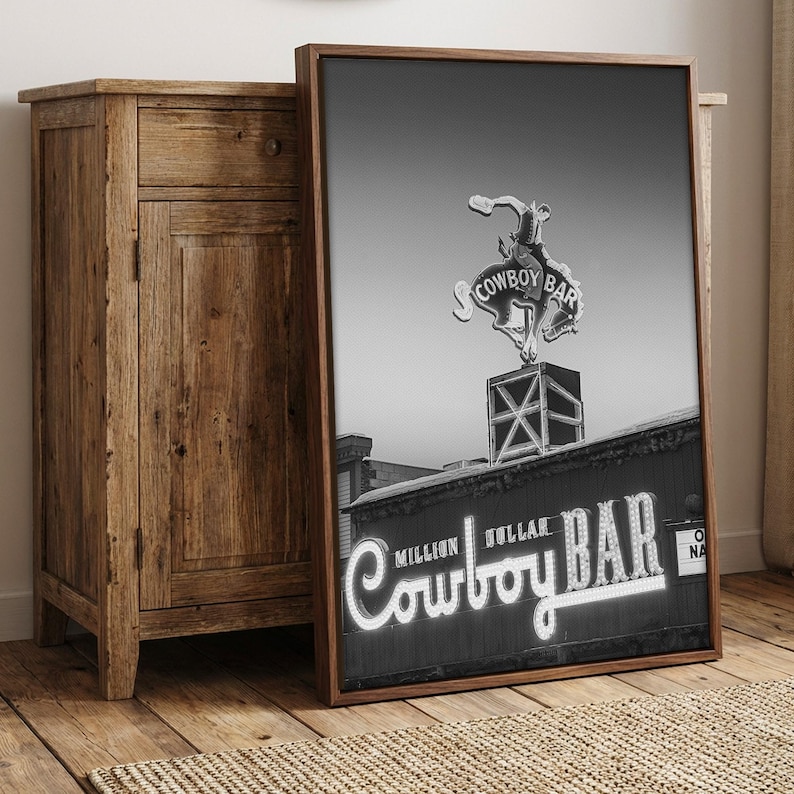 May include: A framed black and white wall art print featuring the Million Dollar Cowboy Bar neon sign. The image displays a bucking bronco rider silhouette atop a box structure, with the text Million Dollar Cowboy Bar in illuminated lettering.