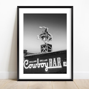 Op de afbeelding: Een zwart-wit ingelijste print met het neonreclamebord van de Million Dollar Cowboy Bar. De afbeelding toont een bokkende bronco-figuur op een vierkante basis, met de tekst Million Dollar Cowboy Bar in grote, cursieve neonletters.