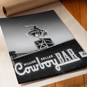 May include: A black and white print featuring the Million Dollar Cowboy Bar sign. The image displays a neon-style sign with the text Million Dollar Cowboy Bar and a silhouette of a bucking bronco rider mounted on a square structure above the roof.