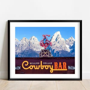 May include: A framed photograph of the Million Dollar Cowboy Bar sign with a neon bucking horse topper against a backdrop of snow-capped mountains. The sign features yellow illuminated lettering and two smaller side signs with text.