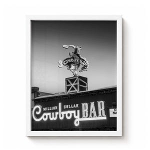 May include: A black and white framed wall art print featuring the Million Dollar Cowboy Bar neon sign. The image displays a bucking bronco figure atop a square base, with the text Million Dollar Cowboy Bar illuminated in script lettering.