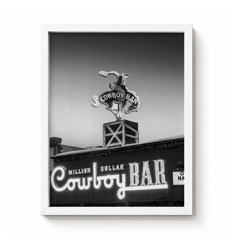 May include: A black and white framed wall art print featuring the Million Dollar Cowboy Bar neon sign. The image displays a bucking bronco figure atop a square base, with the text Million Dollar Cowboy Bar illuminated in script lettering.