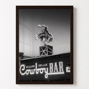 May include: A black and white framed wall art print featuring the Million Dollar Cowboy Bar neon sign. The image displays a bucking bronco cowboy figure atop a box structure, with the text Million Dollar Cowboy Bar illuminated in script.