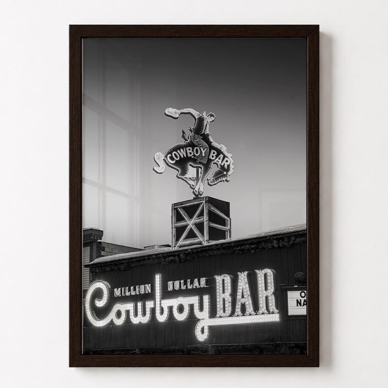 May include: A black and white framed wall art print featuring the Million Dollar Cowboy Bar neon sign. The image displays a bucking bronco cowboy figure atop a box structure, with the text Million Dollar Cowboy Bar illuminated in script.