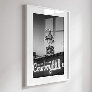 May include: A black and white framed print featuring the Million Dollar Cowboy Bar neon sign. The artwork displays a bucking bronco figure atop a square structure, with the text Million Dollar Cowboy Bar illuminated in a script font style.