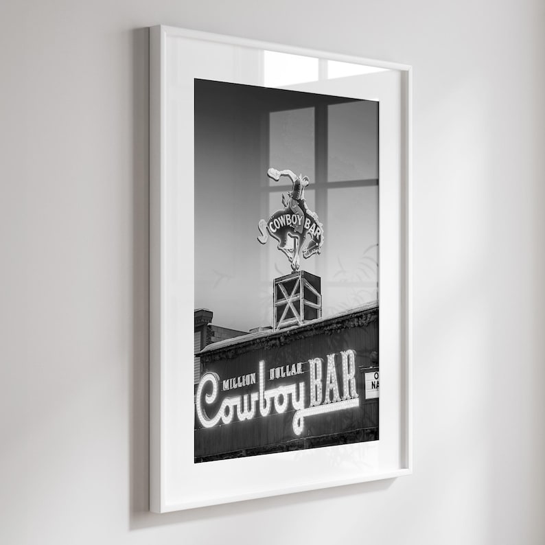 May include: A black and white framed print featuring the Million Dollar Cowboy Bar neon sign. The artwork displays a bucking bronco figure atop a square structure, with the text Million Dollar Cowboy Bar illuminated in a script font style.