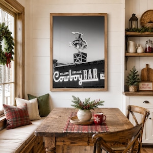 May include: A framed black and white wall art print featuring the Million Dollar Cowboy Bar sign with a bucking horse silhouette. The artwork is displayed in a wooden frame on a white paneled wall above a wooden dining table with holiday decor.