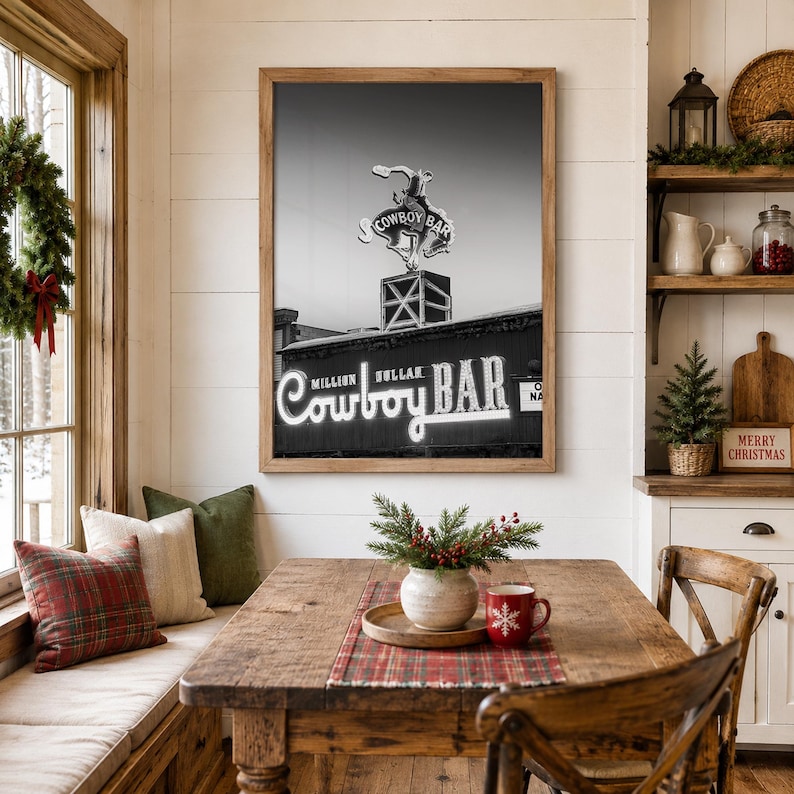 May include: A framed black and white wall art print featuring the Million Dollar Cowboy Bar sign with a bucking horse silhouette. The artwork is displayed in a wooden frame on a white paneled wall above a wooden dining table with holiday decor.