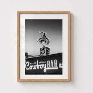 May include: A black and white framed wall art print featuring the Million Dollar Cowboy Bar neon sign. The image displays a bucking bronco silhouette above the text Million Dollar Cowboy Bar in a light-up script font, encased in a light wood frame.