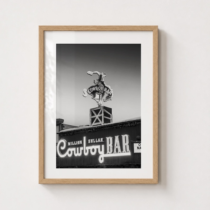 May include: A black and white framed wall art print featuring the Million Dollar Cowboy Bar neon sign. The image displays a bucking bronco silhouette above the text Million Dollar Cowboy Bar in a light-up script font, encased in a light wood frame.