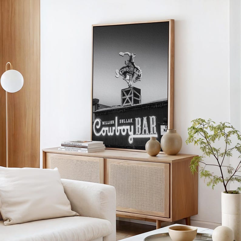 May include: A black and white framed wall art print featuring a neon sign of a bucking bronco above the text Cowboy Bar, with the words Million Dollar Cowboy Bar displayed prominently in a script font at the bottom of the building facade.