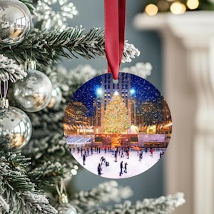 Può includere: Un ornamento natalizio rotondo con una scena festosa di un albero di Natale decorato, pattinatori e palazzi di città di notte. L'ornamento è appeso a un nastro rosso ed è circondato da un albero di Natale innevato.