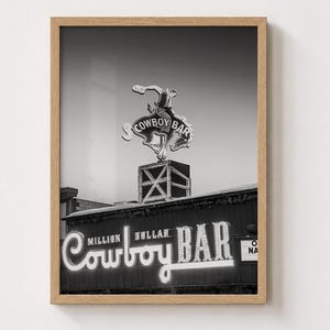 May include: A black and white framed wall art print featuring the Million Dollar Cowboy Bar sign. The image displays a neon sign of a cowboy riding a bucking horse atop a square structure, with the text Million Dollar Cowboy Bar below.