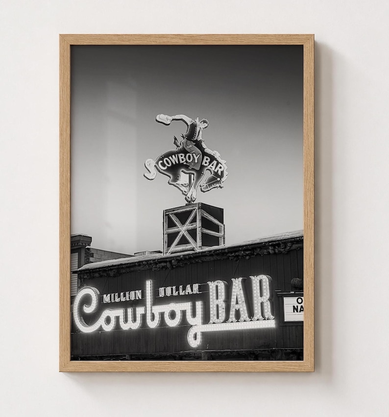 May include: A black and white framed wall art print featuring the Million Dollar Cowboy Bar sign. The image displays a neon sign of a cowboy riding a bucking horse atop a square structure, with the text Million Dollar Cowboy Bar below.
