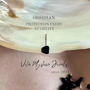 Könnte beinhalten: Ein schwarzer Obsidian-Ohrstecker mit Silberstift vor einem hellrosa strukturierten Hintergrund. Der Text lautet "OBSIDIAN PROTECTION ENERGY STABILITY" und "Vita Mystica Jewels since 2024".