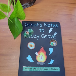 May include: A black notebook with the text "Scout's Notes to Cozy Grove" in white. The cover features colorful illustrations of a car, ghosts, a flame, and other icons. The phrase "All those who are lost deserve kindness" is also on the cover.