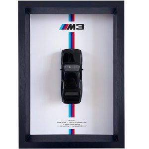 Op de afbeelding: Een zwarte BMW M3 E30 model auto getoond in een zwart frame met een witte achtergrond. De auto staat op een witte streep met blauwe en rode strepen aan beide kanten. De tekst "M3" staat boven de auto en de tekst "M3 E30" staat onder de auto met de volgende specificaties: 286 pk / 182 kW | 2.495 cc 6 cilinders in lijn | 5-versnellingsbak | 0-100 km/h in 6,5 s | Topsnelheid 240 km/h