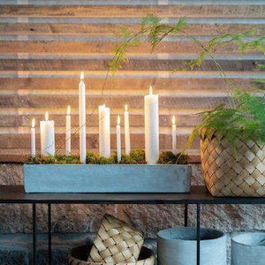 May include: A display of lit white candles in a rectangular concrete planter with moss, on a black metal shelf. Woven baskets and concrete pots are also on the shelf. The background is a wooden wall.