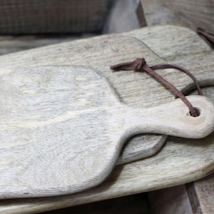May include: Several wooden chopping boards with a pale, distressed finish. The top board features a handle and a leather hanging loop. The boards display a natural wood grain and a rustic appearance.