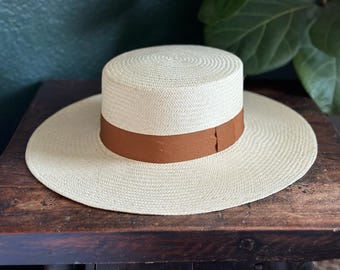 May include: A wide-brimmed straw hat featuring a flat crown and a smooth, solid brown ribbon band encircling the base. The hat is constructed from a tightly woven, cream-colored straw material with a consistent, circular pattern throughout the brim.
