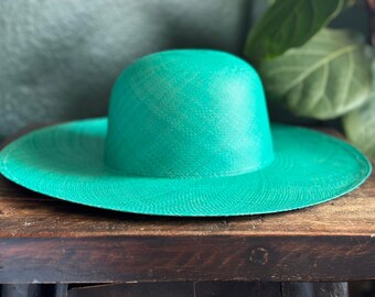May include: A wide-brimmed, turquoise sun hat with a rounded crown. The hat is made of woven material and sits on a dark wooden surface. The hat's brim is approximately 15 inches in diameter, offering ample shade.